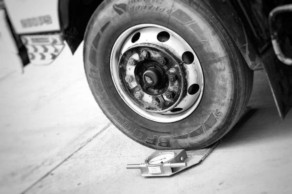Angled close-up of a heavy commercial truck tire resting on a silver portable mechanical wheel scale on a concrete floor to check axle load distribution.
