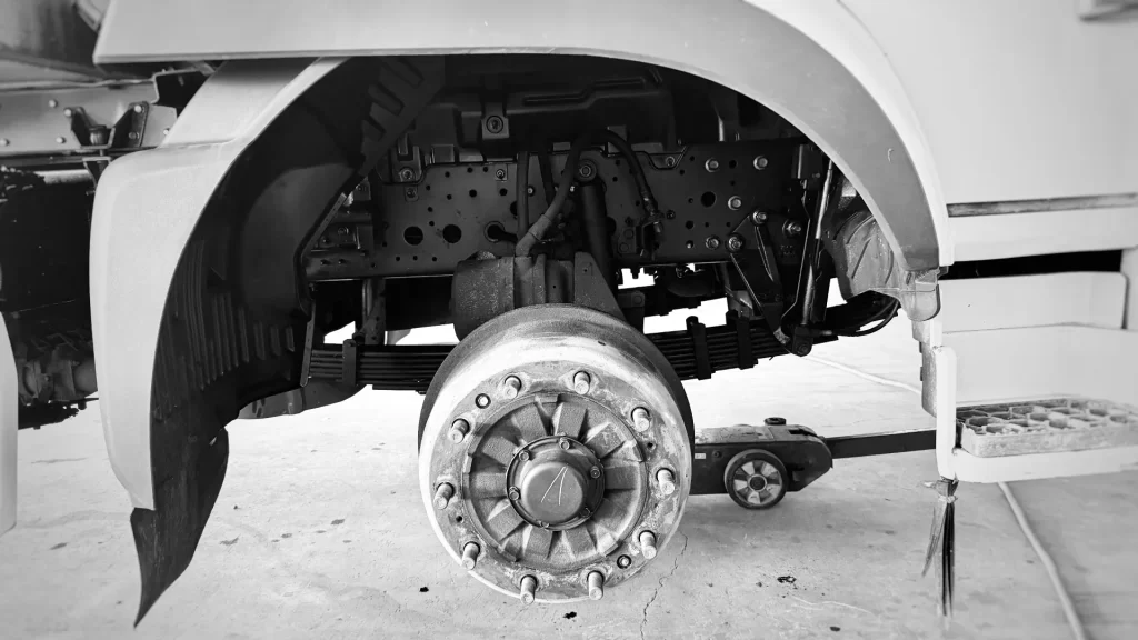 A commercial truck steer axle with the wheel removed, jacked up and prepared for a wheel bearing end-play inspection and suspension component check.